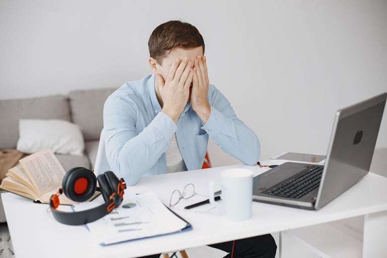 Stressed man at home office desk holding head in frustration over laptop work.