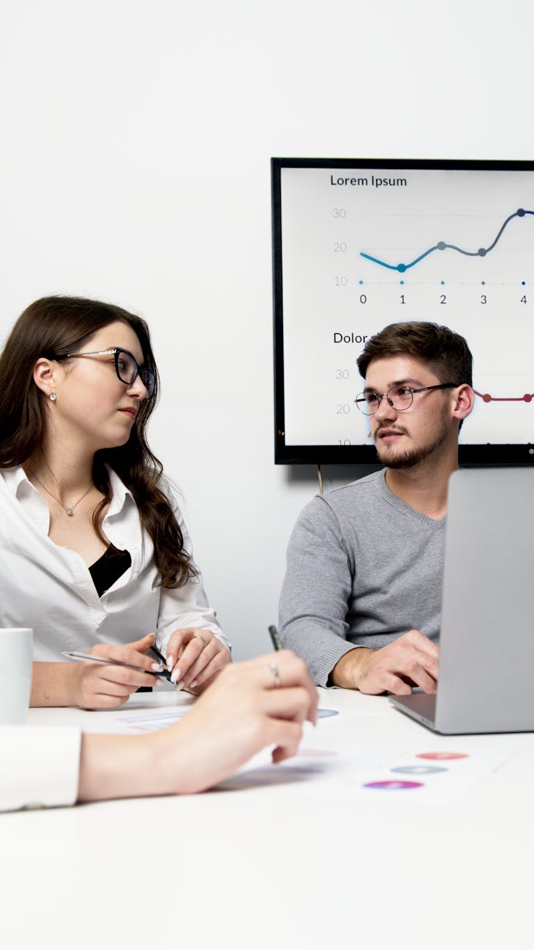 Two colleagues engage in a meeting with data charts displayed on a screen, discussing ideas.